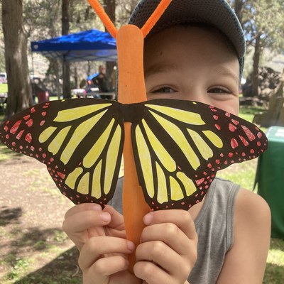 A kiddo holds a yellow and red butterfly craft in front of their smiling face. Photo: Land Trust.